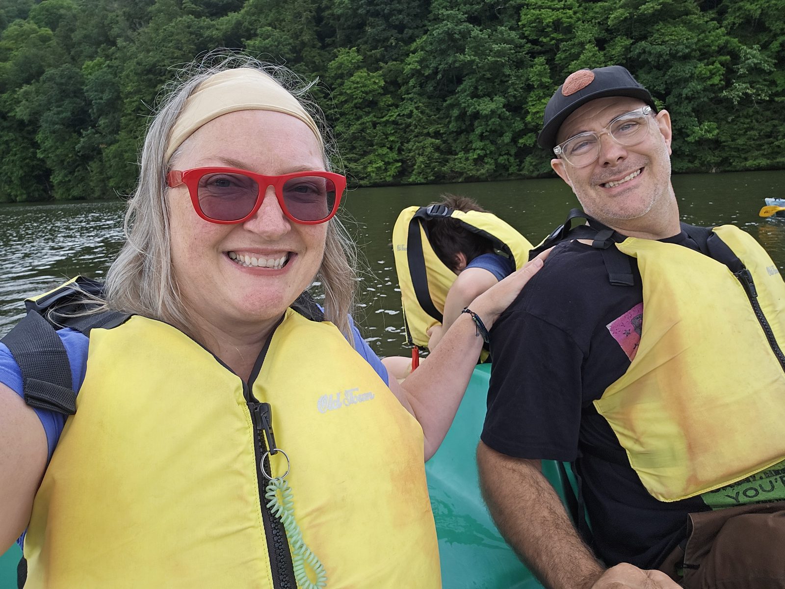 Judy and Adam smiling in yellow life jackets in a kayak on Youghiogheny River Lake near the dam, with a wooded bank behind them